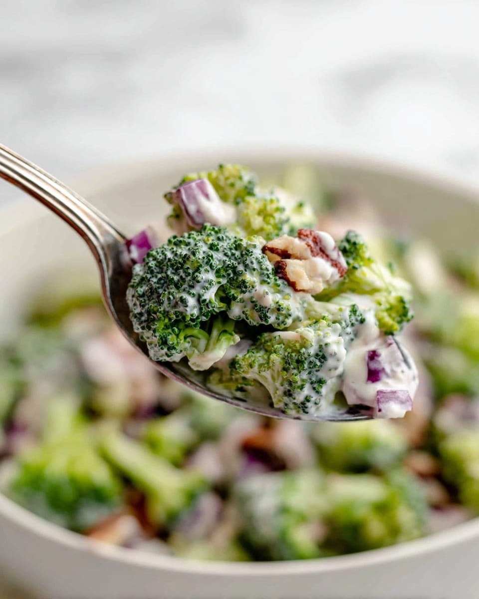 A close-up view of a silver spoon holding a mix of broccoli florets, small light brown nut pieces, and tiny bits of red onion coated with a creamy white dressing, all in front of a blurred white bowl filled with the same broccoli salad. The broccoli is bright green with a slightly rough texture, the nuts add a chewy, irregular shape, and the red onions provide small pops of color. The creamy dressing looks smooth and lightly coats all the ingredients. The background and surface have a white marbled texture, and the scene is softly lit to show freshness. Photo taken with an iphone --ar 4:5 --v 7