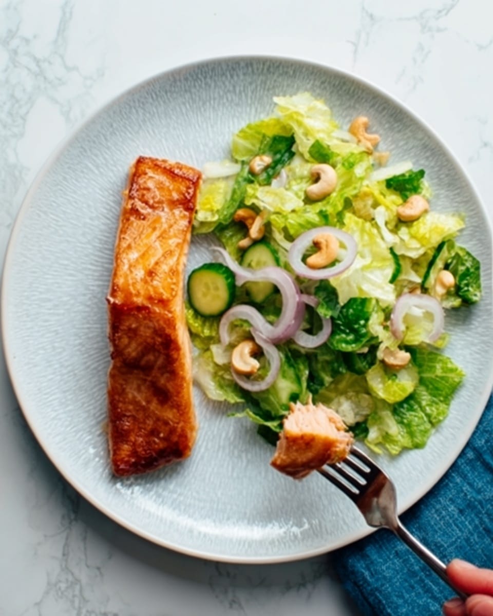 A white round plate sits on a white marbled surface with a single piece of cooked salmon on the left side, its skin crispy and golden brown, showing a textured, flaky appearance. On the right side, there is a fresh salad made of green lettuce pieces mixed with sliced pale green cucumber, thin rings of light purple onion, and white cashew nuts scattered on top. The colors contrast nicely with the warm salmon and cool salad greens. A woman's hand holding a fork with a piece of salmon is visible near the top edge of the plate. photo taken with an iphone --ar 4:5 --v 7