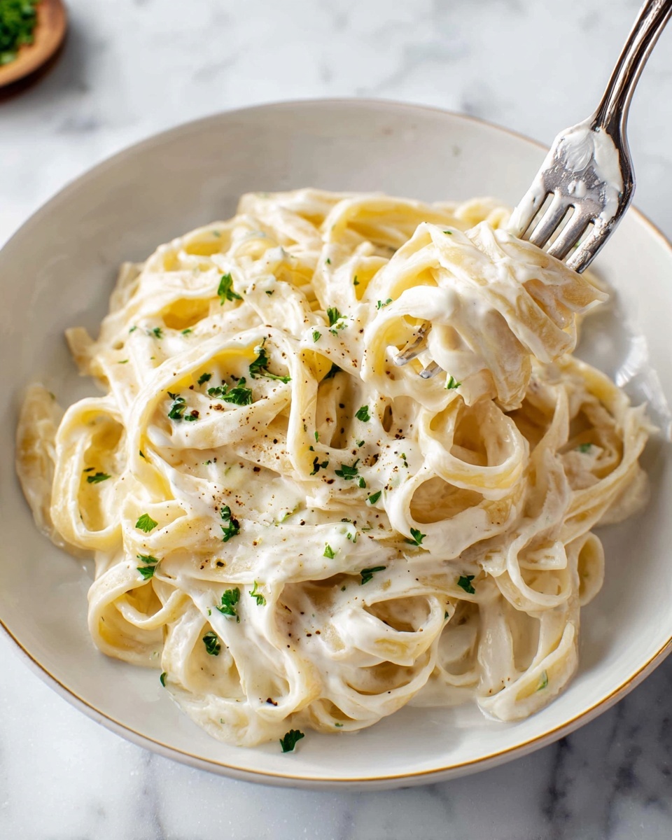 A white bowl holds a single layer of creamy fettuccine pasta coated in thick, pale off-white sauce. The noodles are smooth and slightly twisted, showing their soft texture. Small green parsley pieces are sprinkled evenly on top, adding contrast. A silver fork on the right side lifts a few noodles, showing the sauce’s creamy thickness. The background is a white marbled surface. photo taken with an iphone --ar 4:5 --v 7
