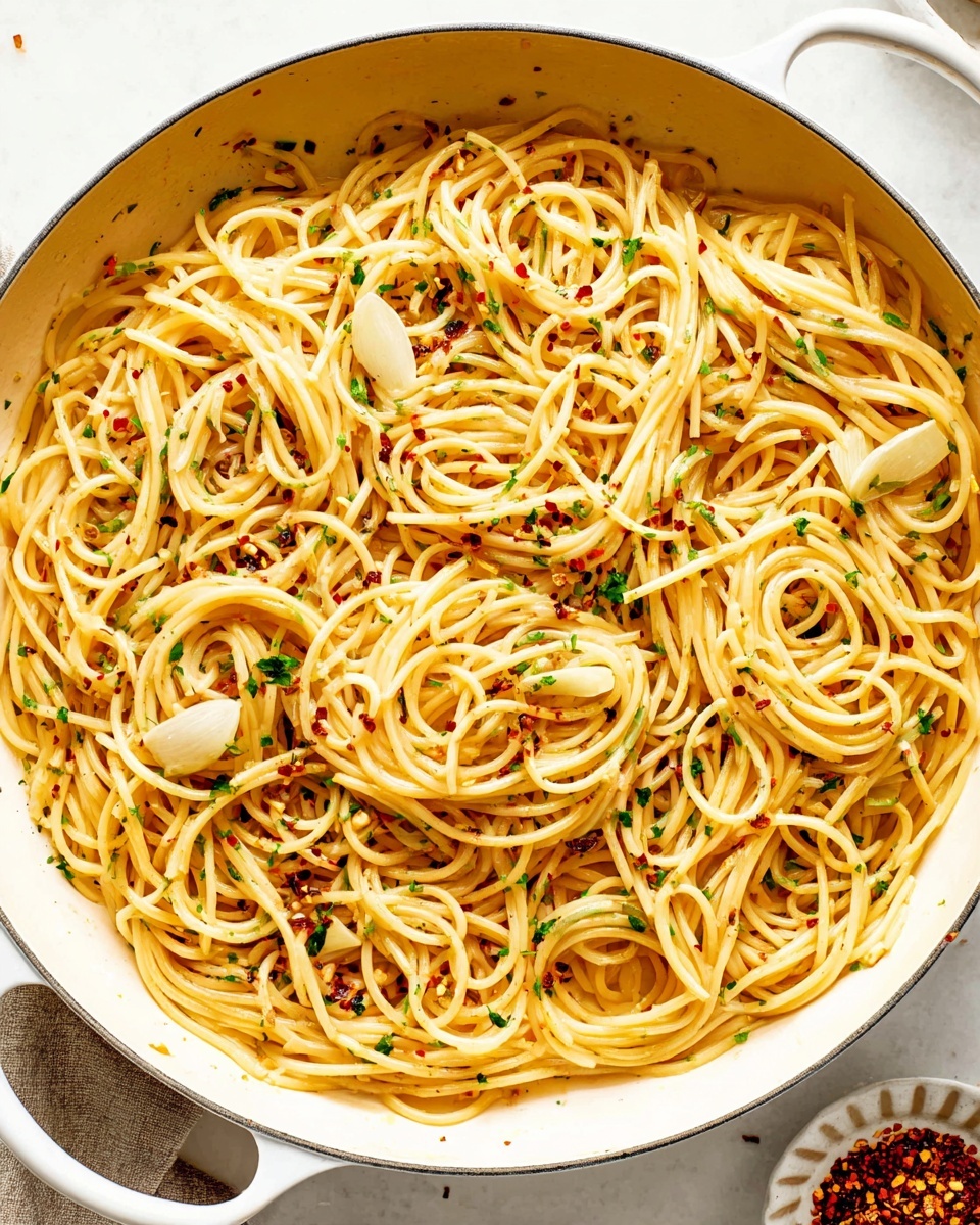 This close-up image shows a plate full of spaghetti pasta. The spaghetti strands are light golden yellow and smooth, twisted and spread all over. Thin slices of light tan garlic are scattered evenly among the pasta, along with small pieces of chopped green parsley and tiny red chili flakes, adding specks of green and red color. The overall look is simple and fresh with a slight shine on the spaghetti from light oil. The background is a white marbled texture. photo taken with an iphone --ar 4:5 --v 7
