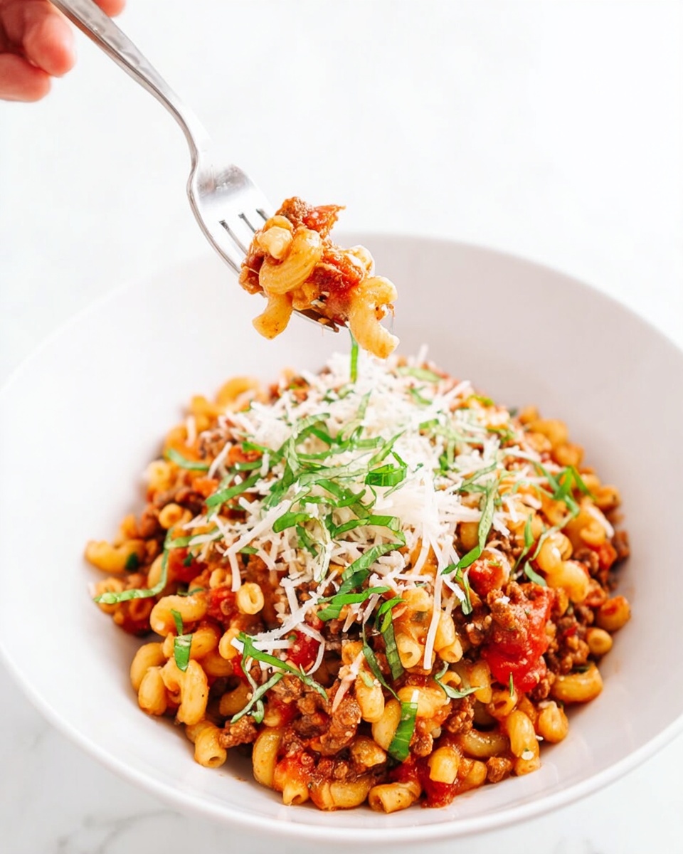 A white plate filled with small curved pasta mixed with chunky red tomato pieces and small bits of browned meat, evenly coated in a reddish sauce. On top, there is a generous layer of finely shredded white cheese sprinkled across the pasta, along with thin strips of fresh green basil scattered lightly. The pasta and sauce mixture forms a slightly heaped mound in the center of the plate. The setting features a white marbled surface in the background. photo taken with an iphone --ar 4:5 --v 7