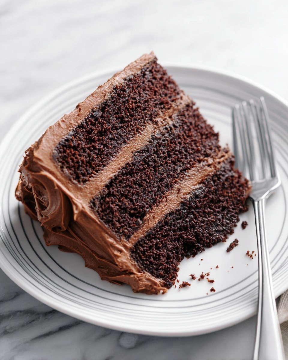 A slice of chocolate cake with three dark brown layers, each separated by smooth light brown chocolate frosting. The entire cake slice is covered on the sides and top by thick, creamy chocolate frosting with visible swirls and texture. The slice is placed on a white plate with thin gray circular lines near its edge, and a fork is resting on the right side of the plate. The setting is on a white marbled texture surface. Photo taken with an iphone --ar 4:5 --v 7