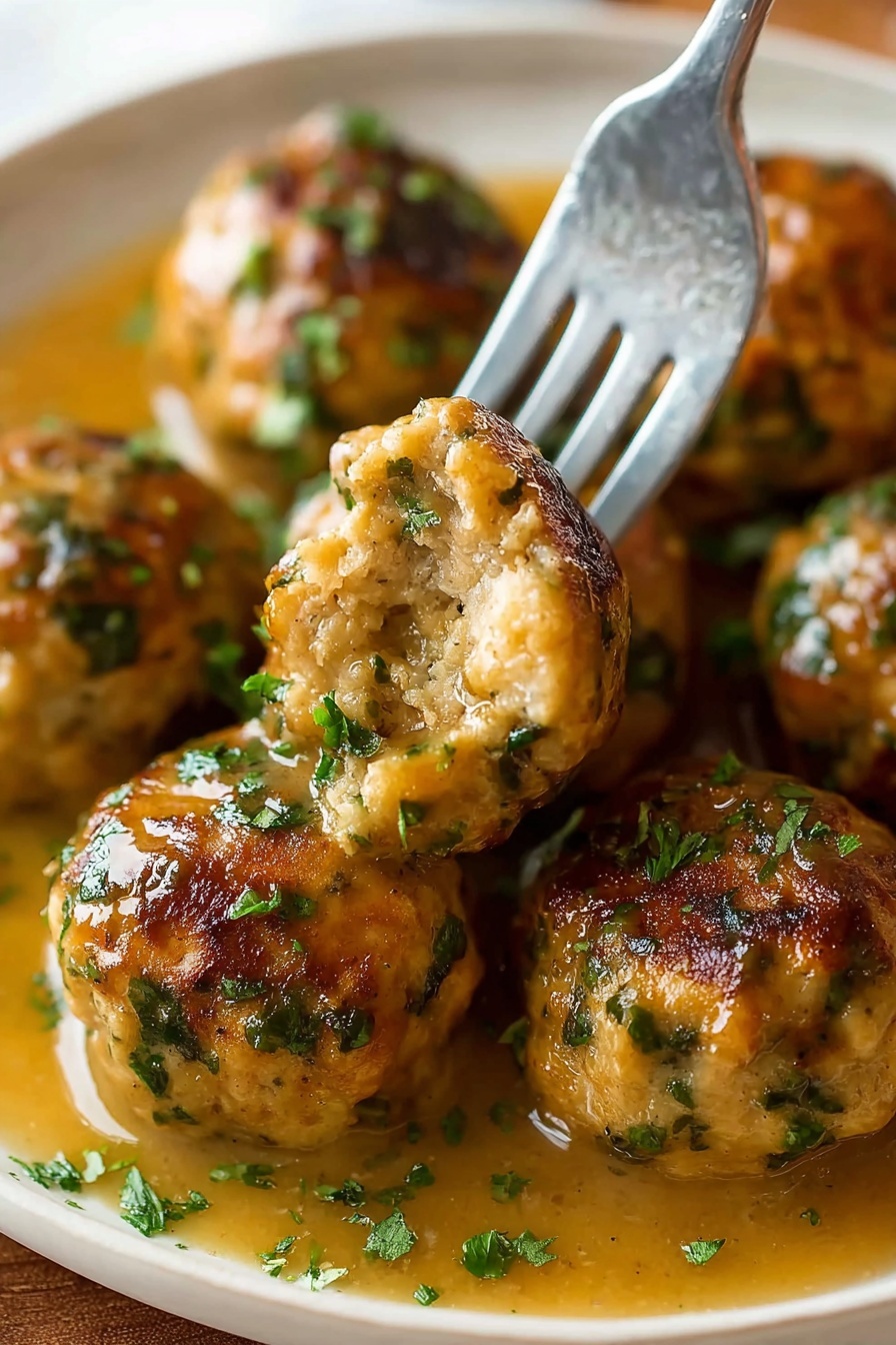 The image shows several round meatballs with a golden-brown crust and green herb bits scattered through them, resting in a light brown gravy. A silver fork holds one meatball with a bite taken out, showing a soft and moist inside with visible herbs. The meatballs are arranged closely on a white plate with some chopped green herbs sprinkled on top and the gravy pooling around them. The background is a white marbled texture photo taken with an iphone --ar 2:3 --v 7 - Duck Fat Roasted Potatoes, crispy roasted potatoes, flavorful potato side dish, golden roasted potatoes, easy duck fat potatoes