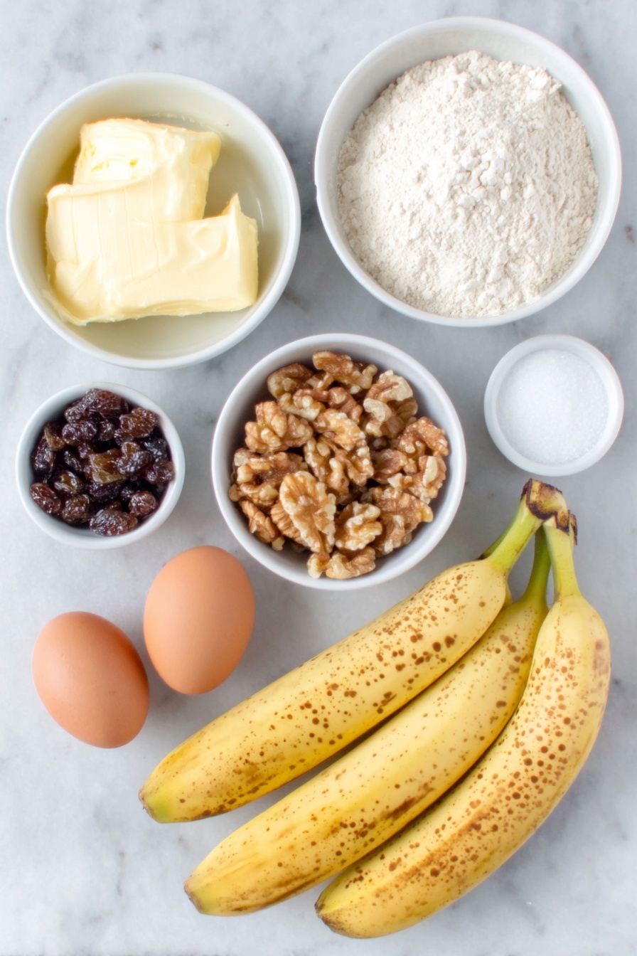 Flat lay of three very ripe bananas with brown speckles, a small white ceramic bowl filled with softened unsalted butter, another white bowl containing granulated sugar, two whole uncracked brown eggs, a white bowl with all-purpose flour, a small white bowl holding walnuts roughly chopped, a small white bowl filled with dark raisins, a small white bowl with pale beige baking soda powder, a tiny white bowl with coarse salt, and a small white bowl with golden vanilla extract, all arranged with perfect symmetry on a clean white marble surface, soft natural light, photo taken with an iPhone, professional food photography style, fresh ingredients, white ceramic bowls, no bottles, no duplicates, no utensils, no packaging --ar 2:3 --v 7 --p m7354615311229779997 - Moist Banana Bread with Walnuts and Raisins, banana bread with walnuts and raisins, easy banana bread recipe, homemade banana bread, healthy banana bread with nuts and fruits