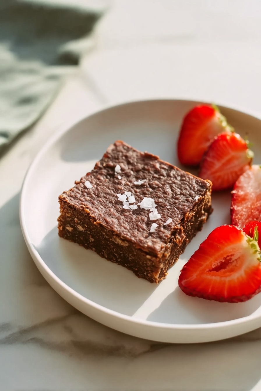 A white round plate sits on a white marbled surface holding a single square piece of dark brown dessert with a rough, textured top sprinkled lightly with flaky white salt. Next to the dessert are two halves of a bright red strawberry, one showing the inner juicy flesh and the other showing the dotted exterior and green leafy top. The scene focuses mainly on the plate with soft natural light highlighting the colors and textures. photo taken with an iphone --ar 2:3 --v 7 - Chocolate Banana Baked Oatmeal, healthy baked oatmeal with chocolate and bananas, easy breakfast ideas, nutritious chocolate banana oats, wholesome breakfast recipes