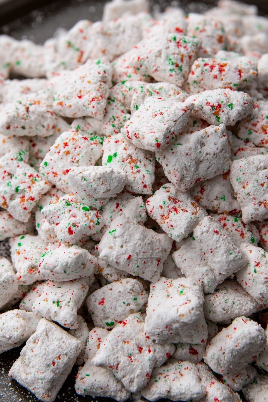 This image shows a large pile of small, irregularly shaped white pieces covered in a powdery coating. The white coating has specks of red and green sprinkled over all the pieces, giving a festive look. The pieces have a rough and uneven texture, suggesting they might be crunchy or dry. They are spread out on a dark tray, but the main focus is the texture and color of the coated pieces. The red and green sprinkles are small and scattered evenly across the surface of each piece, adding a colorful contrast to the white powder coating. photo taken with an iphone --ar 2:3 --v 7 - Christmas Puppy Chow, festive puppy chow, holiday snack recipes, chocolate peanut butter treats, Christmas party snacks