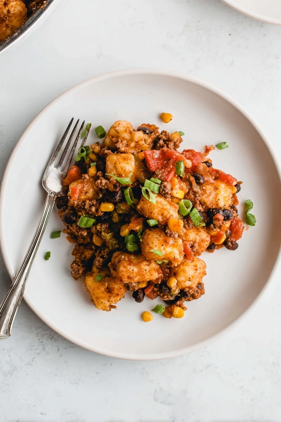 A white plate sits on a white marbled surface, holding a layered dish. The bottom layer includes browned, small potato puffs that are crispy. Mixed in are bits of yellow corn and black beans, creating specks of yellow and black. There are pieces of green onions scattered on top, adding a fresh green color. Red tomato pieces and small bits of a brown sauce or meat mixture cover the dish, giving it a rich texture. A silver fork rests on the left side of the plate partly under the food. Photo taken with an iphone --ar 2:3 --v 7 - Cowboy Casserole with Tater Tots, easy cowboy casserole, cheesy tater tot casserole, hearty family casseroles, comfort food recipes
