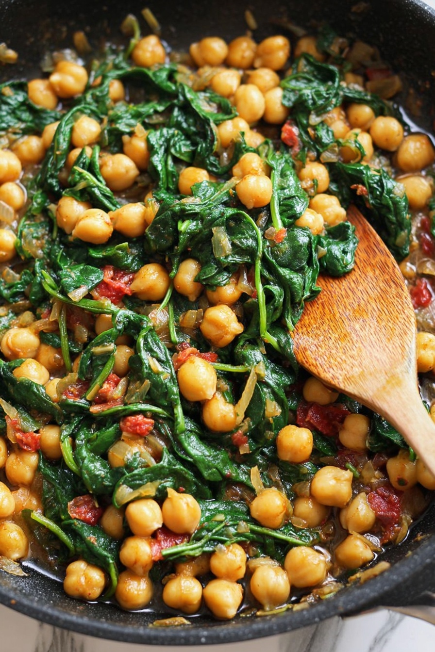 A close-up view of a black skillet filled with cooked chickpeas and wilted dark green spinach leaves mixed with small pieces of light yellow onions and bits of red tomato, producing a rich combination of yellow, green, and red colors. A wooden spoon rests on the right side of the skillet, partially submerged in the mixture, and the background shows a white marbled texture. The overall look is warm, colorful, and rustic, highlighting the fresh ingredients and their cooked textures. photo taken with an iphone --ar 2:3 --v 7 - Spicy Chickpeas with Tomatoes and Spinach, spicy chickpeas, healthy chickpea dishes, quick vegan dinners, flavorful vegetarian recipes