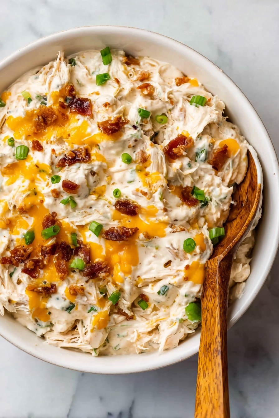 The image shows a white bowl filled with a creamy dip that has a thick, chunky texture. The dip contains visible strands of shredded white meat mixed with creamy white sauce. There are small bright orange melted cheese drops spread over the top and small pieces of green onion scattered throughout. Crispy brown bits, likely bacon or a similar garnish, are also mixed in, adding texture. A wooden spoon with a smooth surface rests inside the bowl on the right side. The bowl is placed on a white marbled surface. photo taken with an iphone --ar 2:3 --v 7 - Crock Pot Crack Chicken, easy slow cooker chicken recipes, creamy cheesy bacon chicken, family-friendly Crock Pot meals, simple slow cooker dinner ideas
