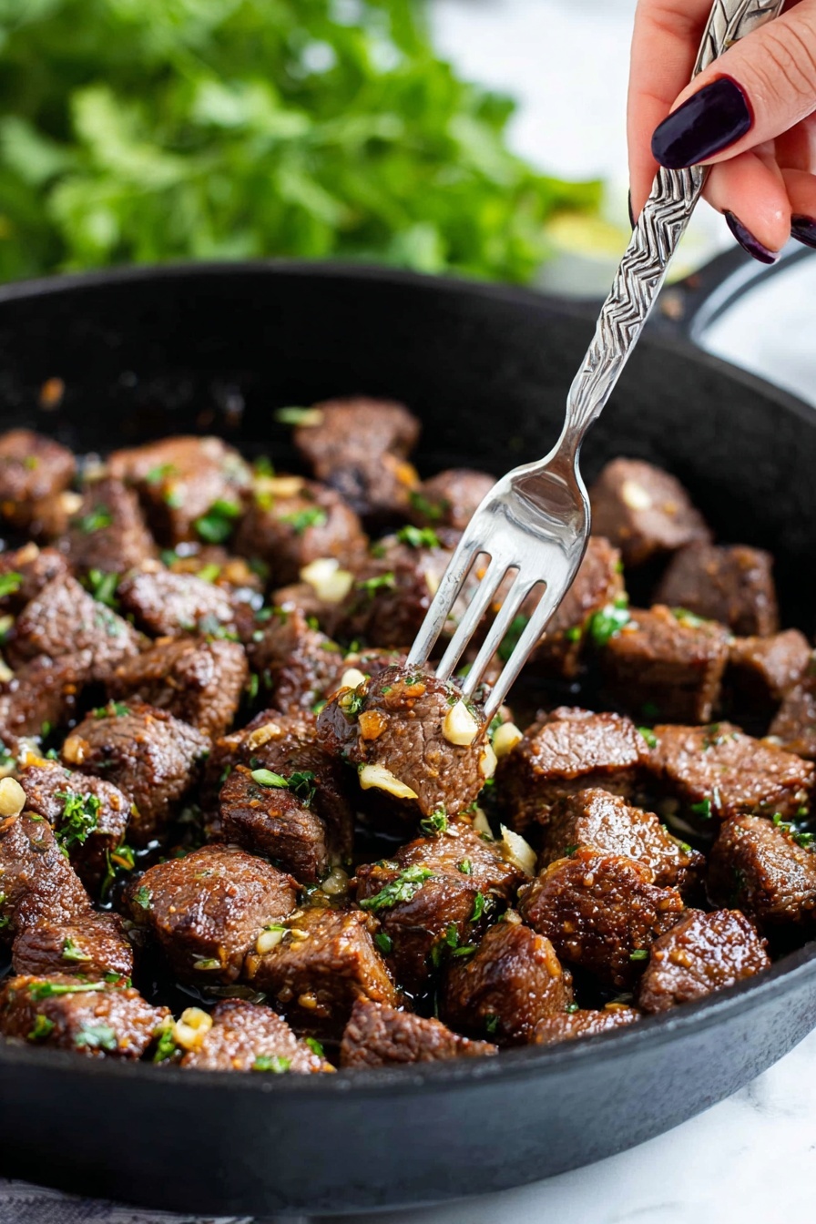 The image shows a black cast iron pan filled with many small chunks of cooked brown meat, each piece sprinkled with bits of green herbs and small pieces of golden-brown garlic. A woman's hand with dark nail polish is holding a silver fork that picks up a single piece of meat from the pan. The background is blurry with green leaves visible, creating a fresh feel. The pan sits on a white marbled surface. photo taken with an iphone --ar 2:3 --v 7 - Garlic Butter Steak Bites, garlic butter steak bites, easy steak bites, tender steak bites, flavorful steak appetizer