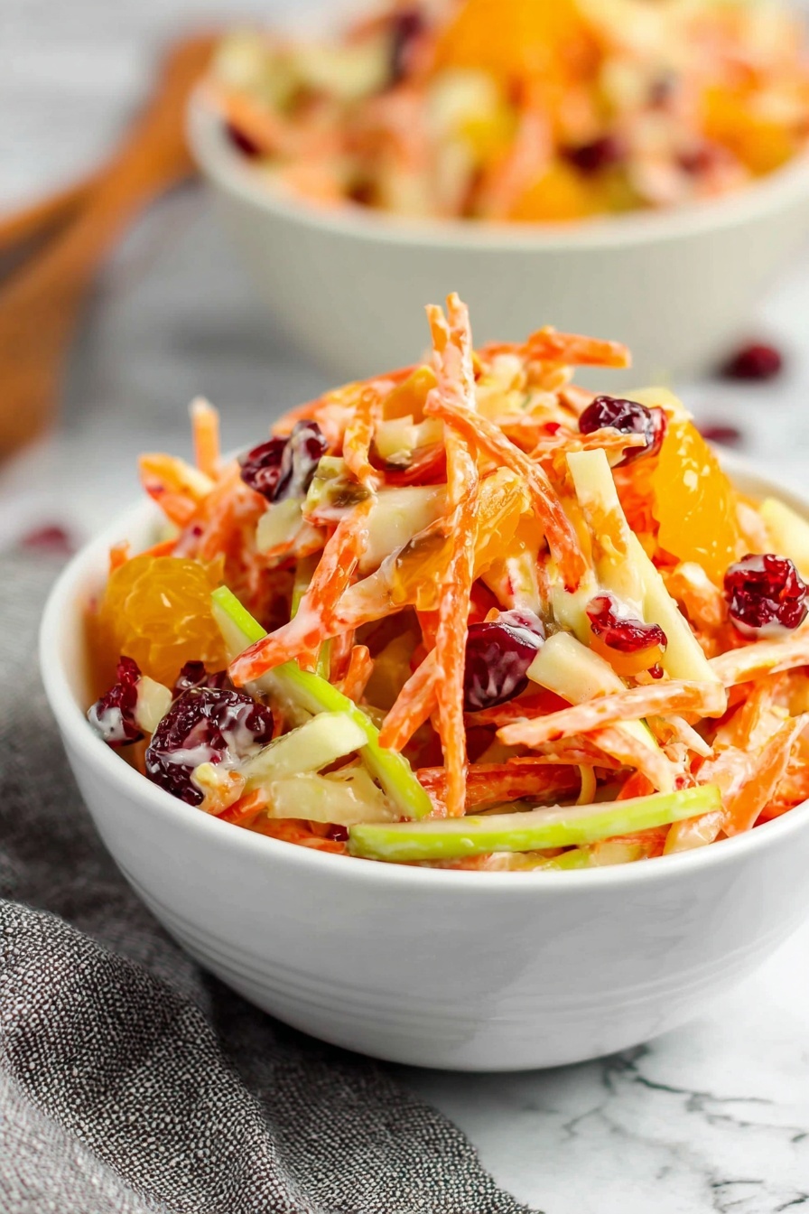 A white bowl is filled with a colorful salad that has shredded orange carrots, thin slices of green apple, small chunks of white apple, bright orange mandarin segments, and dark red dried cranberries mixed together. The salad looks creamy with a light dressing coating the pieces. The bowl sits on a white marbled surface with a gray cloth slightly blurred in the background. Another bowl with the same salad is partly visible behind the main bowl. photo taken with an iphone --ar 2:3 --v 7 - Carrot Apple Salad with Cranberries and Mandarin, healthy fruit salad, fresh vegetable fruit salad, easy colorful salad recipe, tangy sweet fruit salad