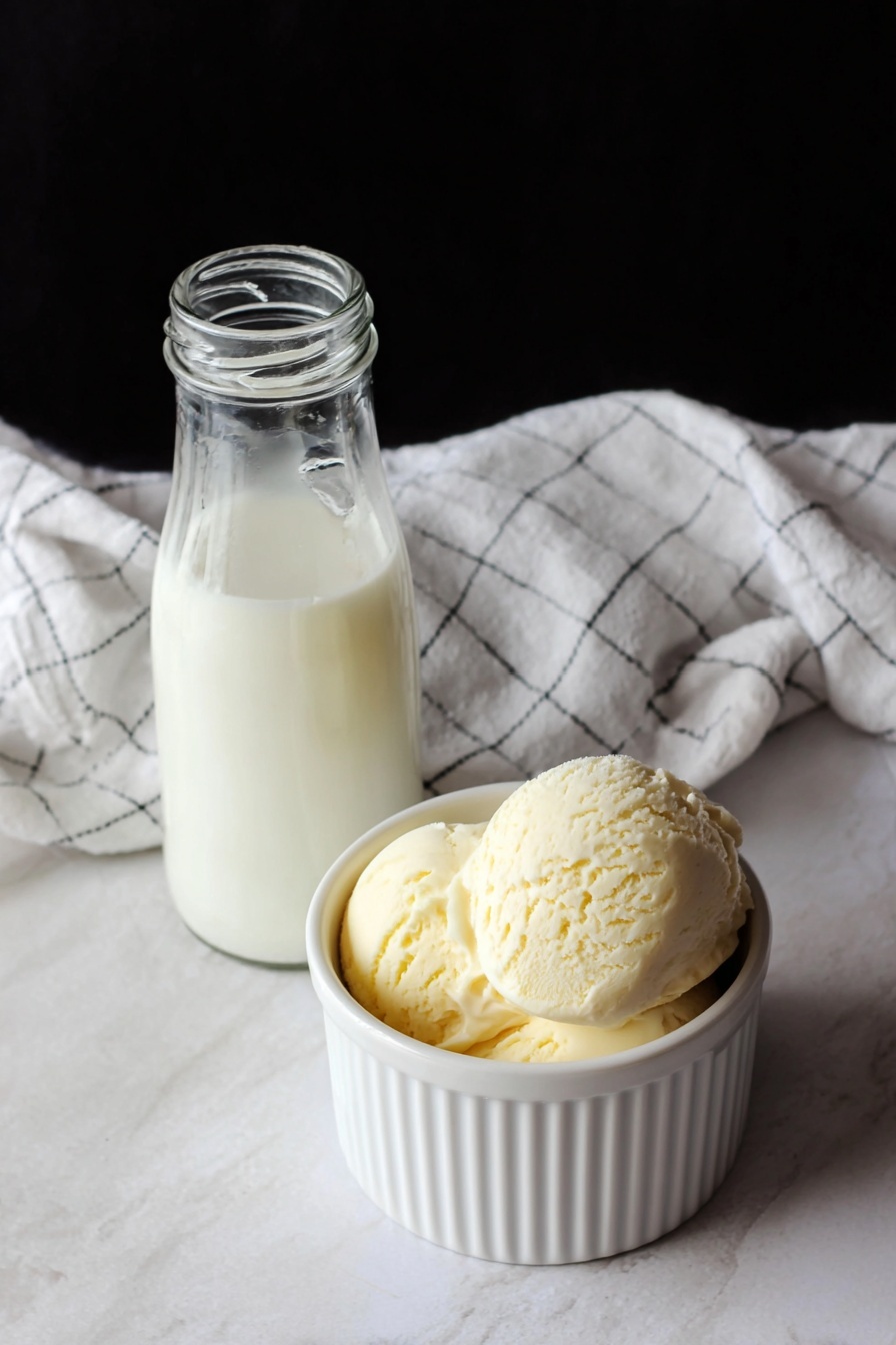 The image shows two scoops of pale yellow ice cream in a small white ribbed ceramic bowl placed on a white marbled surface. The ice cream has a smooth texture with some rough edges on top of the scoops, giving it a creamy and soft look. In the background, slightly out of focus, is a small glass bottle filled with milk, adding a simple and clean touch to the scene. The background is black, which contrasts with the white bowl and the ice cream. photo taken with an iphone --ar 2:3 --v 7 - Homemade Sweet Cream Ice Cream, vanilla ice cream, DIY ice cream, creamy homemade ice cream, easy ice cream recipe