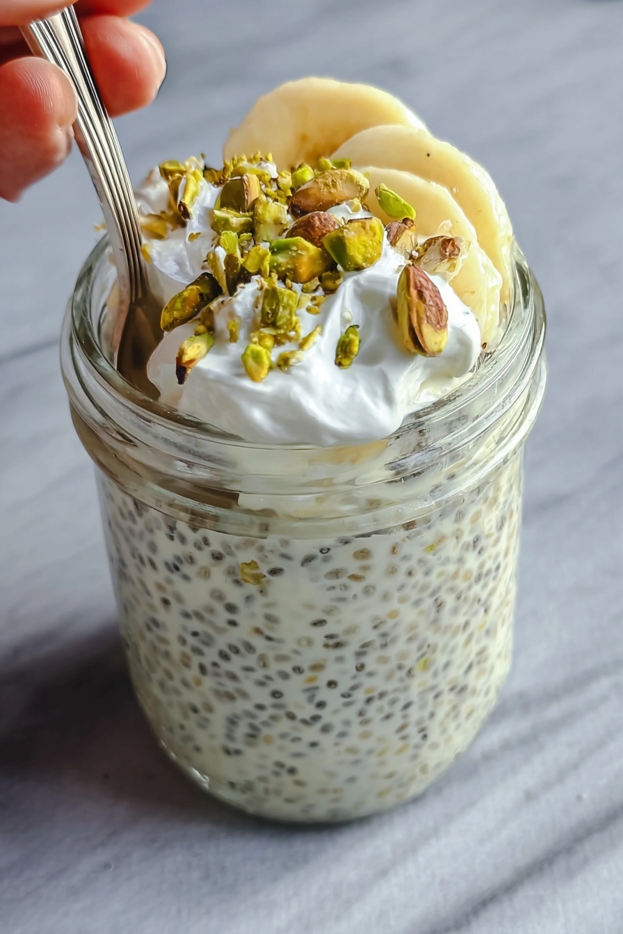 The image shows a clear glass jar filled almost to the top with a creamy white chia pudding that has small black chia seeds spread evenly throughout. On the top layer inside the jar, there are a few slices of light yellow banana, which are partially covered by the creamy pudding. Over the bananas, there is a layer of small white whipped cream dollops, adding a soft texture. Scattered on the very top are roughly chopped green pistachio nuts that add a contrasting color and crunchy texture. A spoon is placed inside the jar, with a woman's hand holding it gently from the left side. The jar is set against a smooth, white marbled surface. Photo taken with an iphone --ar 2:3 --v 7 - Creamy Pistachio Protein Overnight Oats, pistachio overnight oats, healthy protein breakfast, easy overnight oats recipe, high protein morning meal