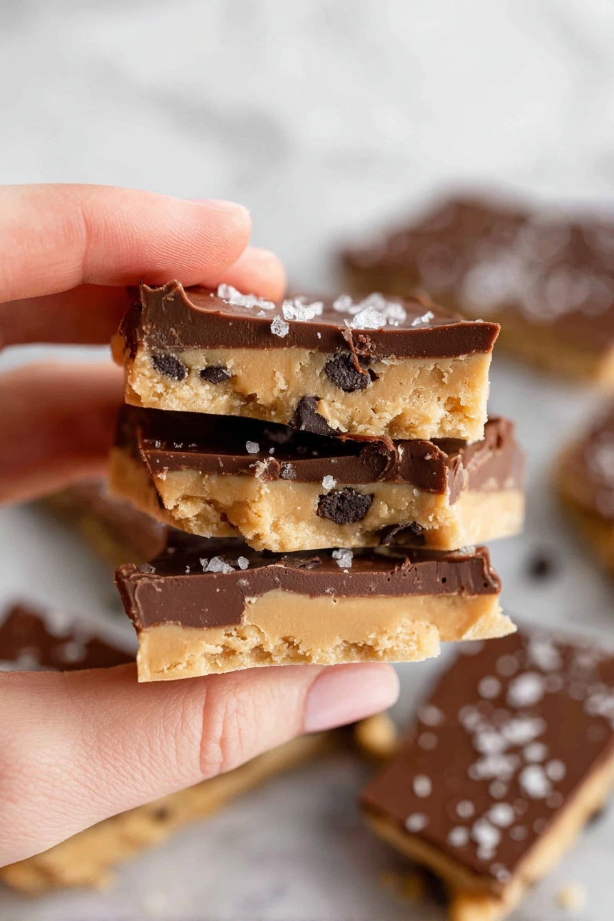 A woman's hand is holding three pieces of snack bark stacked closely together. Each piece has two layers: a bottom light tan layer with a soft texture and dark chocolate chip bits mixed in, and a thick top layer of smooth, dark brown chocolate. The top chocolate layer is sprinkled with small flakes of white sea salt. More broken pieces of the same snack are scattered on a white marbled surface in the background, showing both tan and dark chocolate layers clearly. Photo taken with an iphone --ar 2:3 --v 7 - Healthy Cookie Dough Bark, healthy cookie dough treats, guilt-free cookie dough snack, wholesome dessert ideas, no-bake healthy sweets