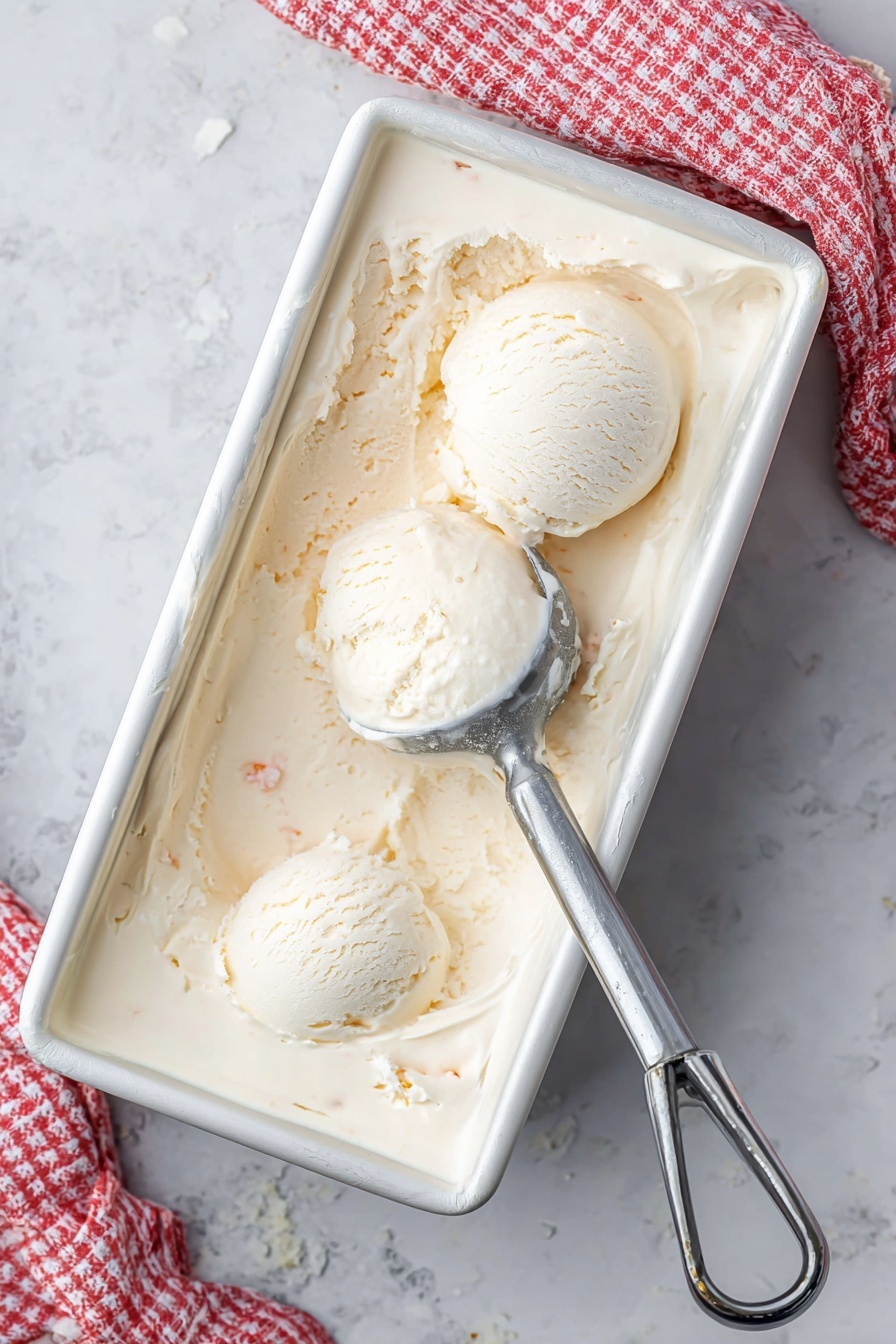 A white metal rectangular container filled with creamy, pale beige ice cream shows two round scoops placed near the top left corner. Ice cream has a smooth but slightly textured surface with small visible bits inside. A silver ice cream scooper rests partially buried on the right side, with some ice cream residue on it. A white and red checkered cloth is partly visible in the upper right corner on a white marbled surface. photo taken with an iphone --ar 2:3 --v 7 - Homemade Coconut Ice Cream, coconut ice cream, tropical ice cream, dairy-free coconut dessert, creamy coconut ice cream